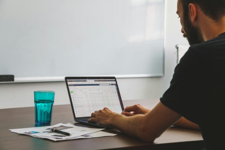 man working at desk on a laptop with a glass of water and some paperwork in front of a white board