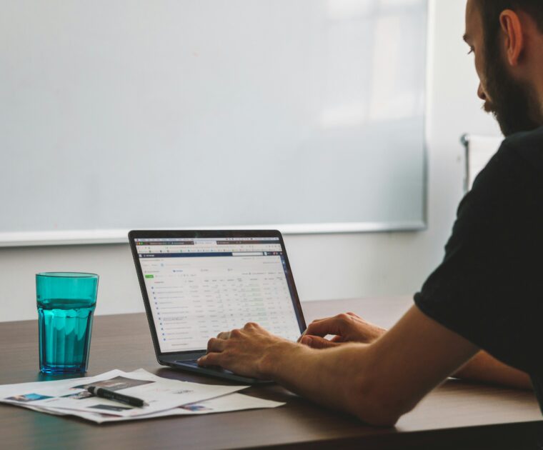 man working at desk on a laptop with a glass of water and some paperwork in front of a white board